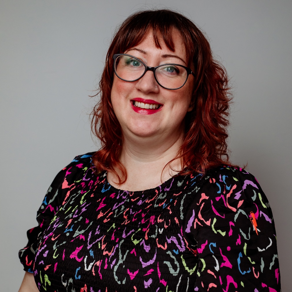 This is a portrait of Sam Taylor, Digital Learning Team Lead at Catalyst IT Europe. Sam is smiling and wearing a colourful, patterned blouse with hues of pink, purple, green, and blue against a black background. They have glasses, and their hair is styled in loose waves. The background is neutral