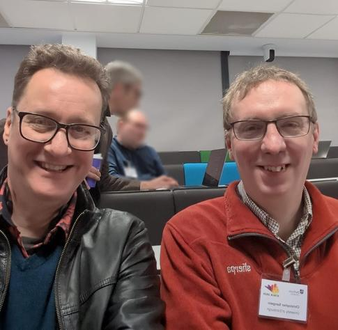 A photo of two men sitting side by side, smiling at the camera. On the left is Marcus Green from Catalyst IT Europe, wearing a black leather jacket and glasses. On the right is Professor Chris Sangwyn, creator of STACK, wearing a red fleece jacket with "copsite" written on it. Both are in a classroom setting.