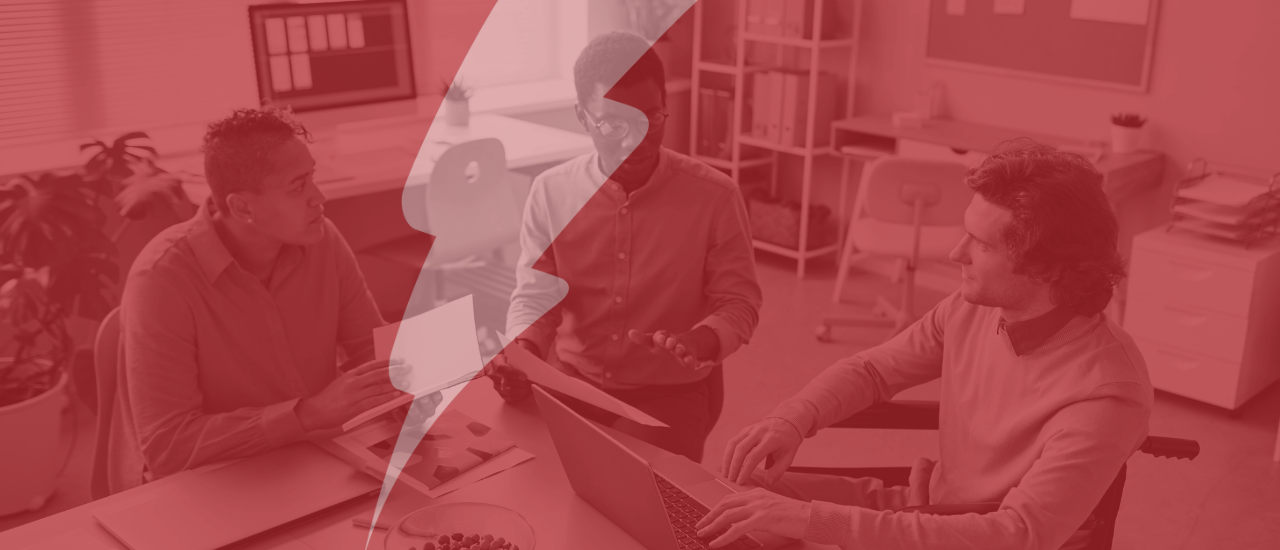 Three colleagues sitting around a table in an office space, having a discussion, with one person holding papers and another working on a laptop. The image is overlaid with a red filter and a transparent lightning bolt graphic in the centre, symbolising energy or impact.