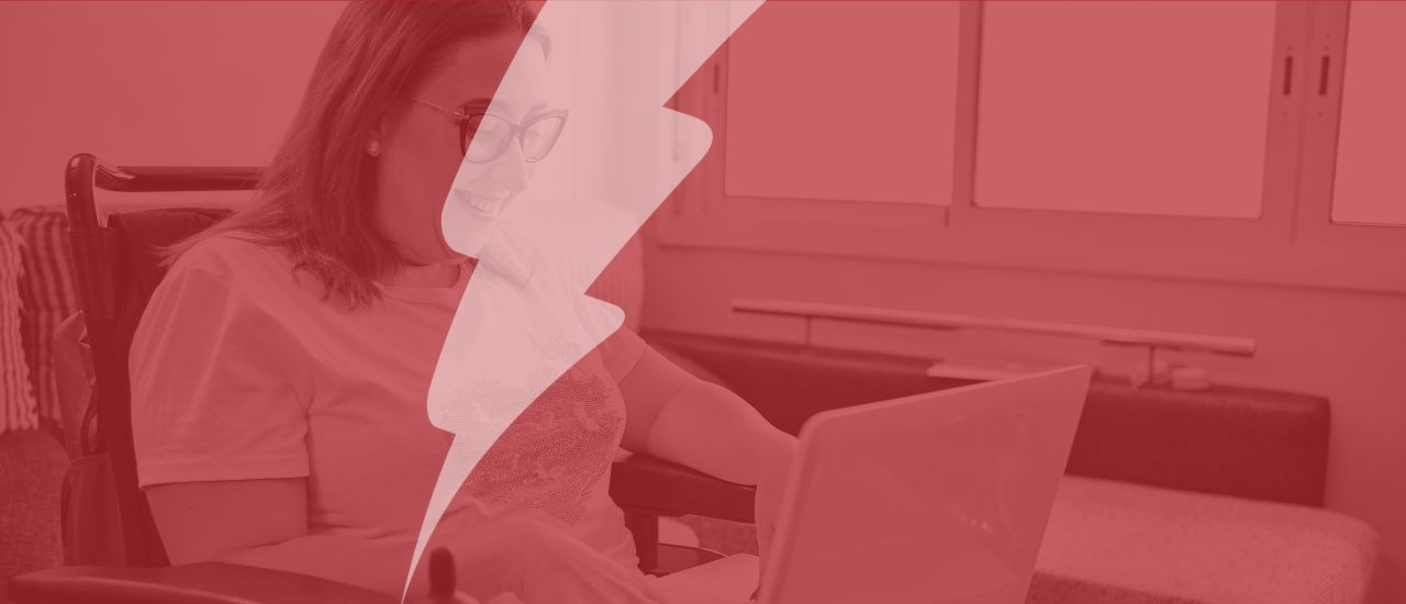 Person sitting in a wheelchair smiling while using a laptop at home, with a sofa and large windows in the background. The image is overlaid with a red filter and a bold lightning bolt graphic running through the centre, symbolising energy and transformation in digital accessibility.