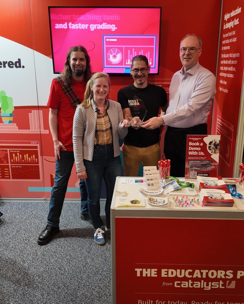Group photo taken at the Catalyst IT stand during MoodleMoot Global 2025. From left to right: Mark Johnson (Catalyst IT Europe), Antonia Bonaccorso (ETH Zürich), Luca Bösch (Bern University of Applied Sciences), and Tim Hunt (The Open University, UK). They are smiling and holding the Community Contributor Award in front of a red display showcasing the Educators Pack by Catalyst. The booth features promotional materials, demo signage, and a screen displaying analytics graphics with the tagline “Richer teaching tools and faster grading.